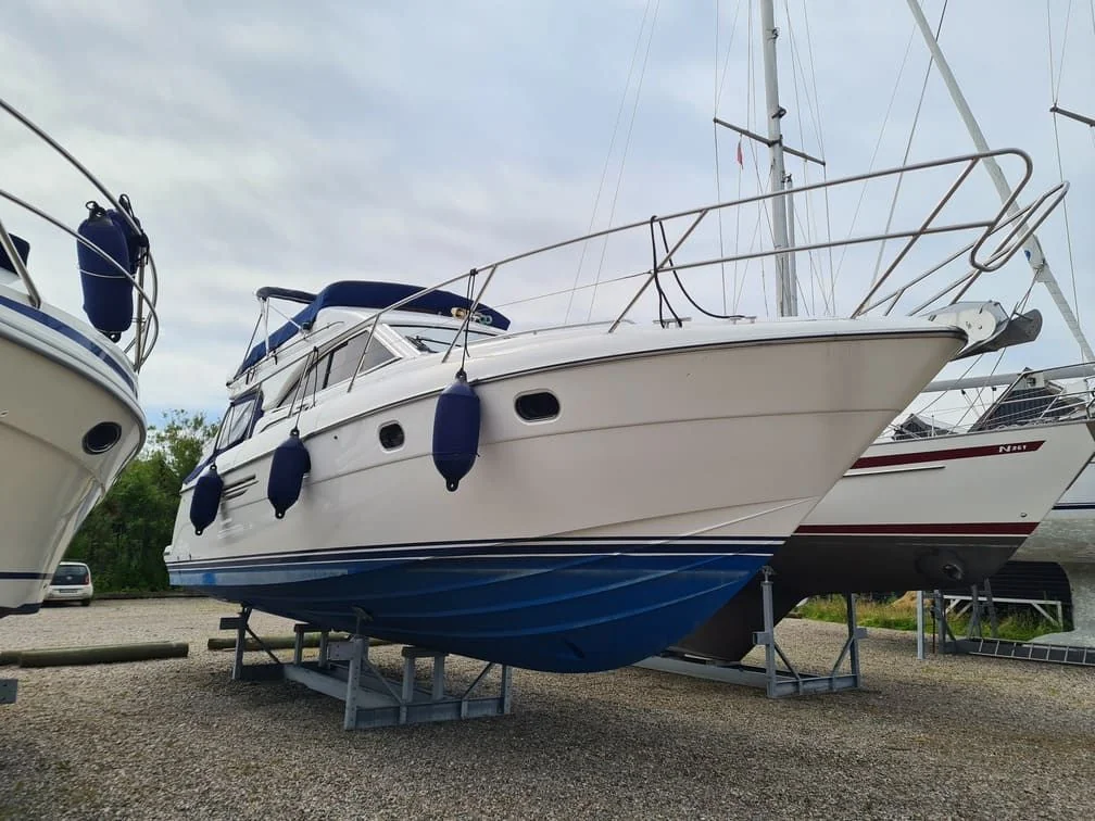 Cabin cruiser sailboat on trailer in storage yard with rigging and fenders visible.