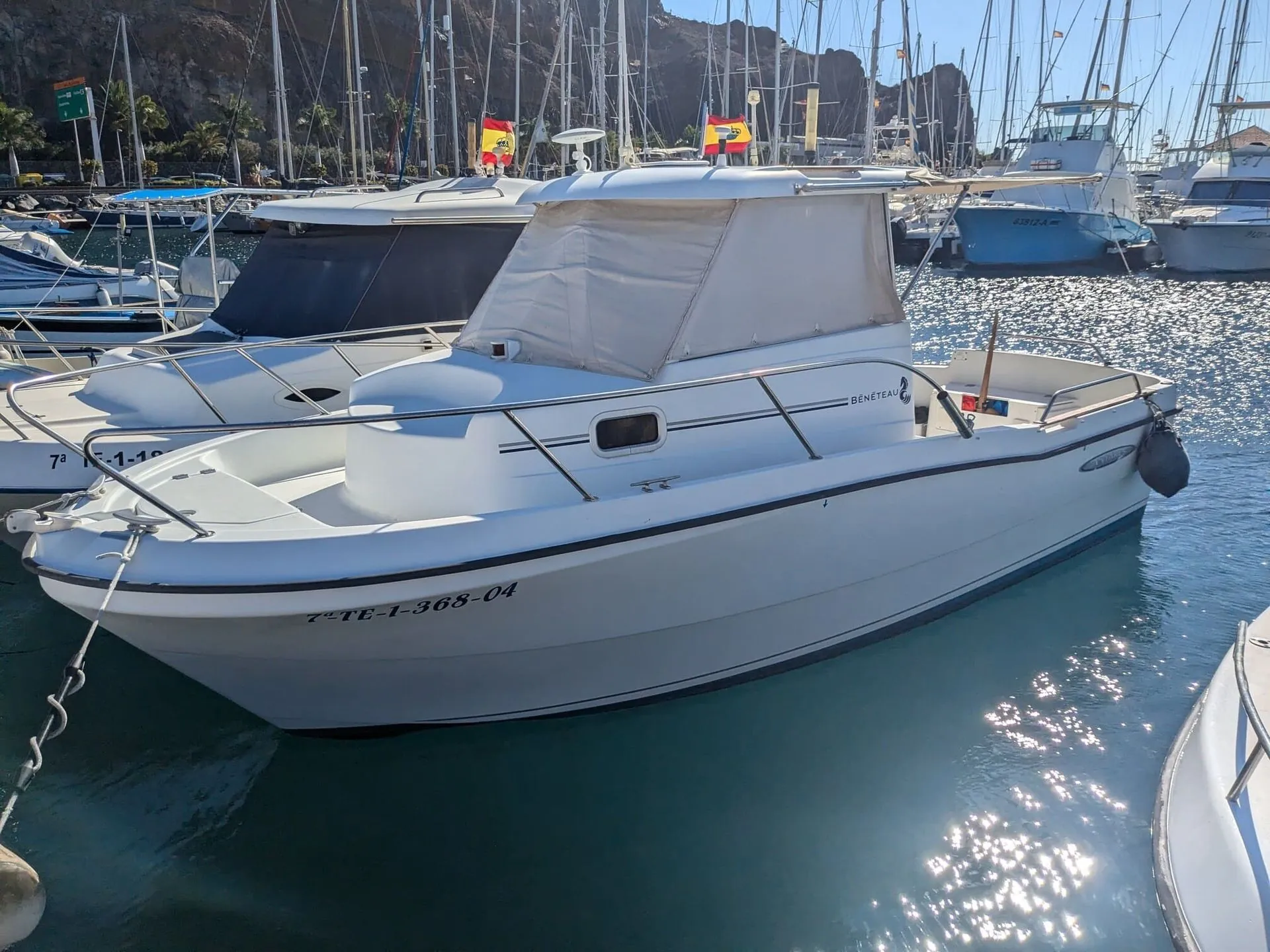 White fiberglass motorboat moored in Mediterranean marina with sailboats and rocky cliffs.
