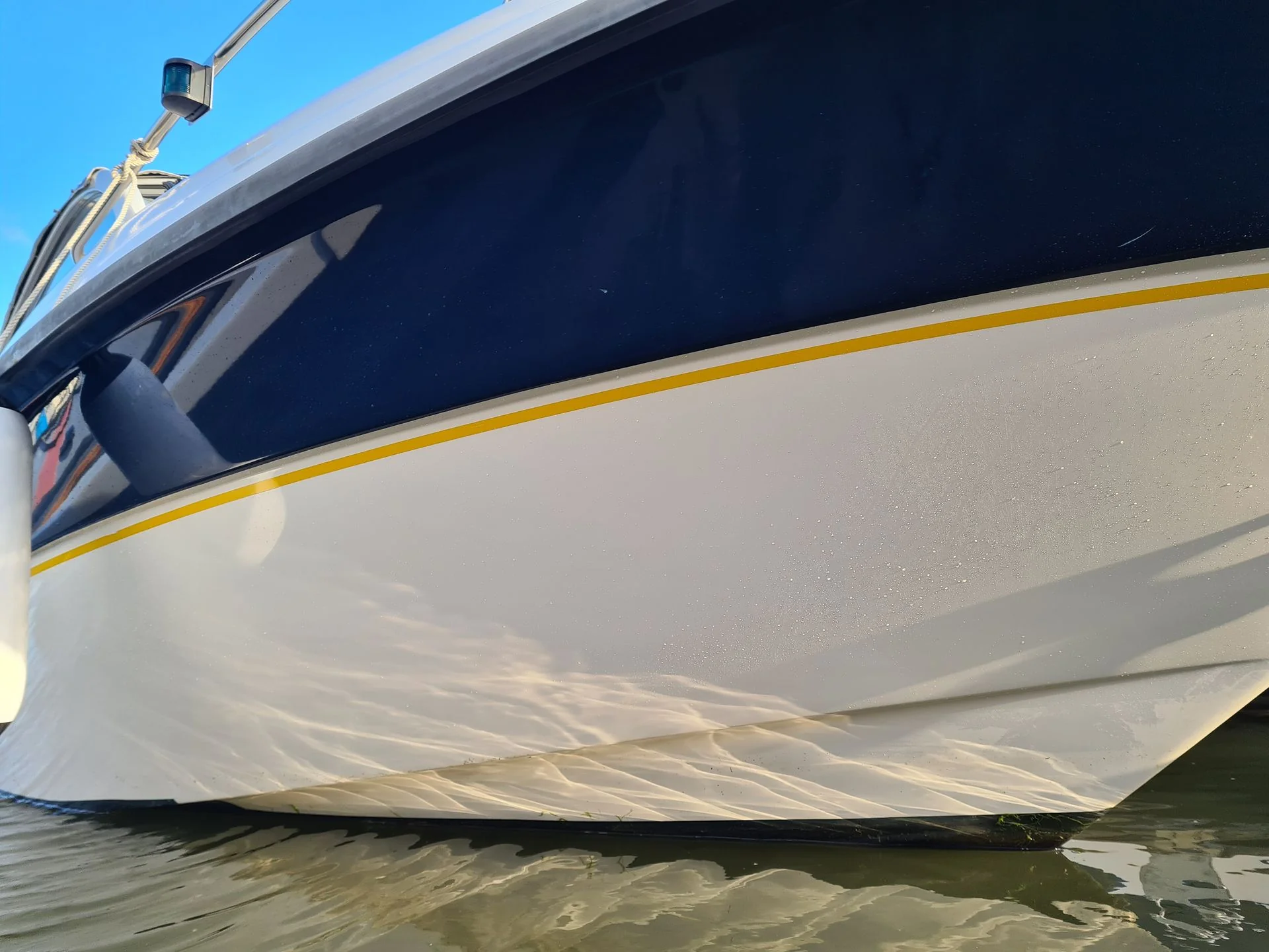 Fiberglass boat hull with yellow stripe detail in shallow water, showing weathered gelcoat surface.