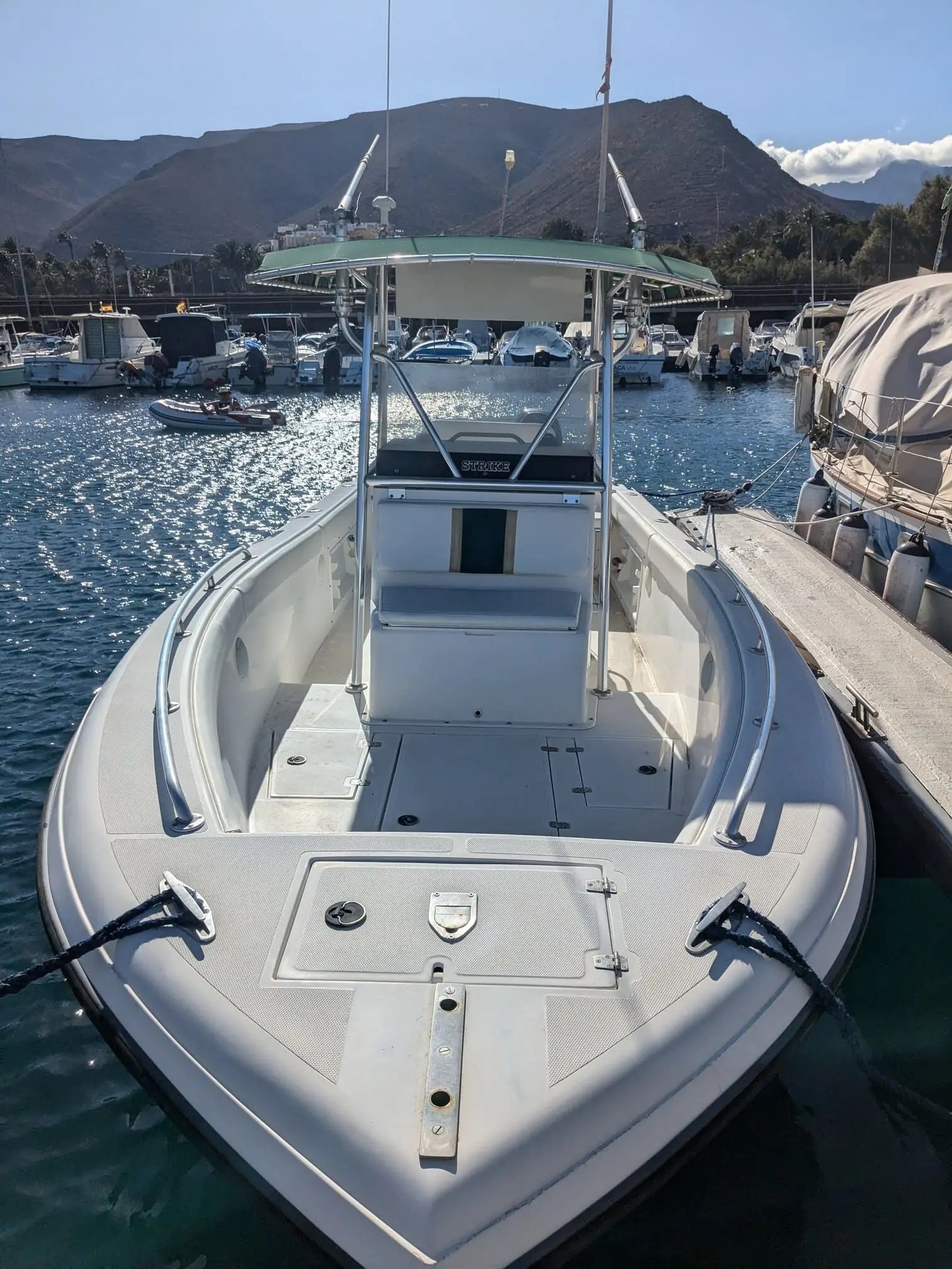 White fiberglass motorboat docked at marina with outboard engine visible, surrounded by other vessels and mountainous backdrop.